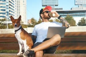 Young Man With Beard Tattoos Laptop His Knees Is Drinking Coffee From Paper Cup His Dog Sits Him